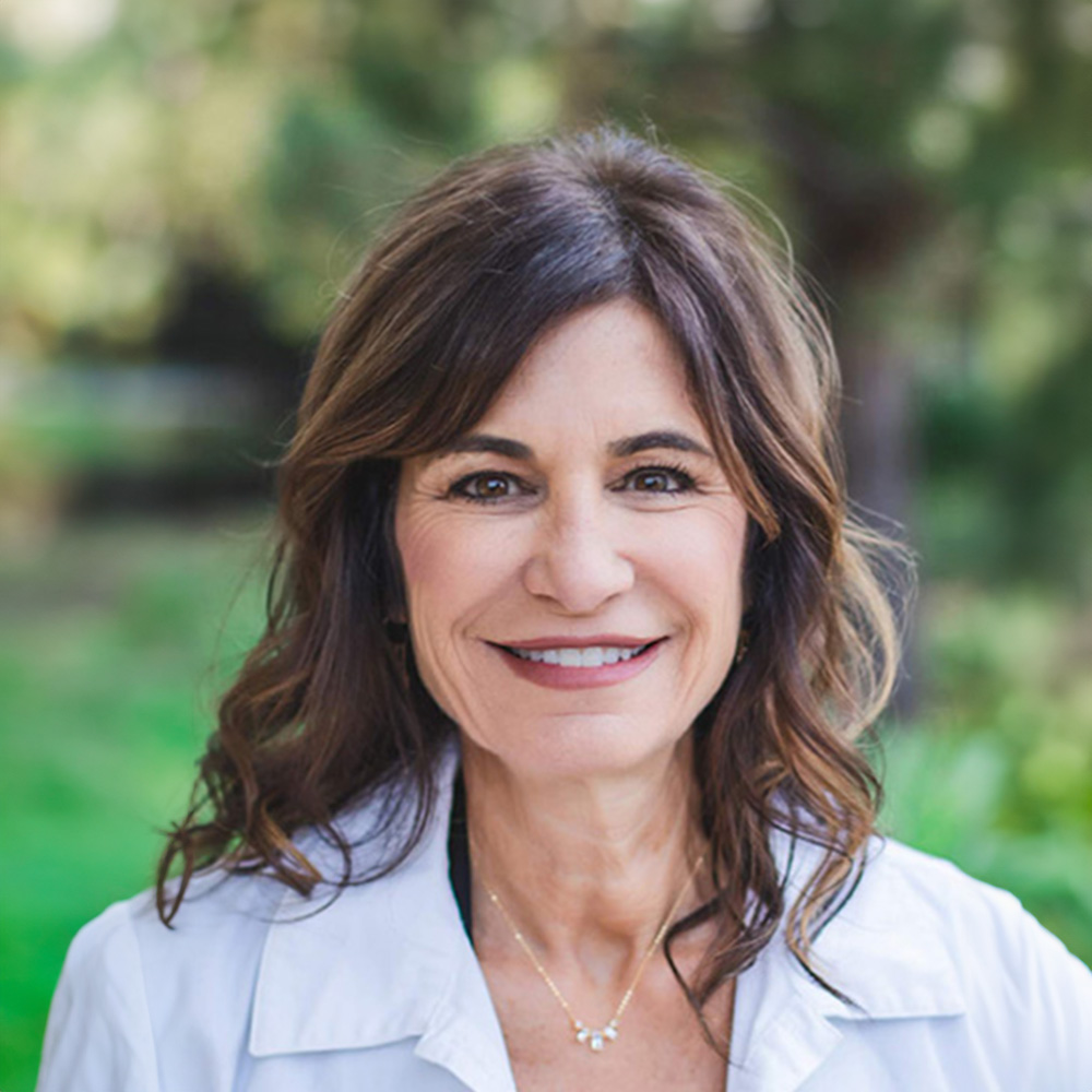 A smiling woman with short hair wearing a white jacket stands against a blurred background.