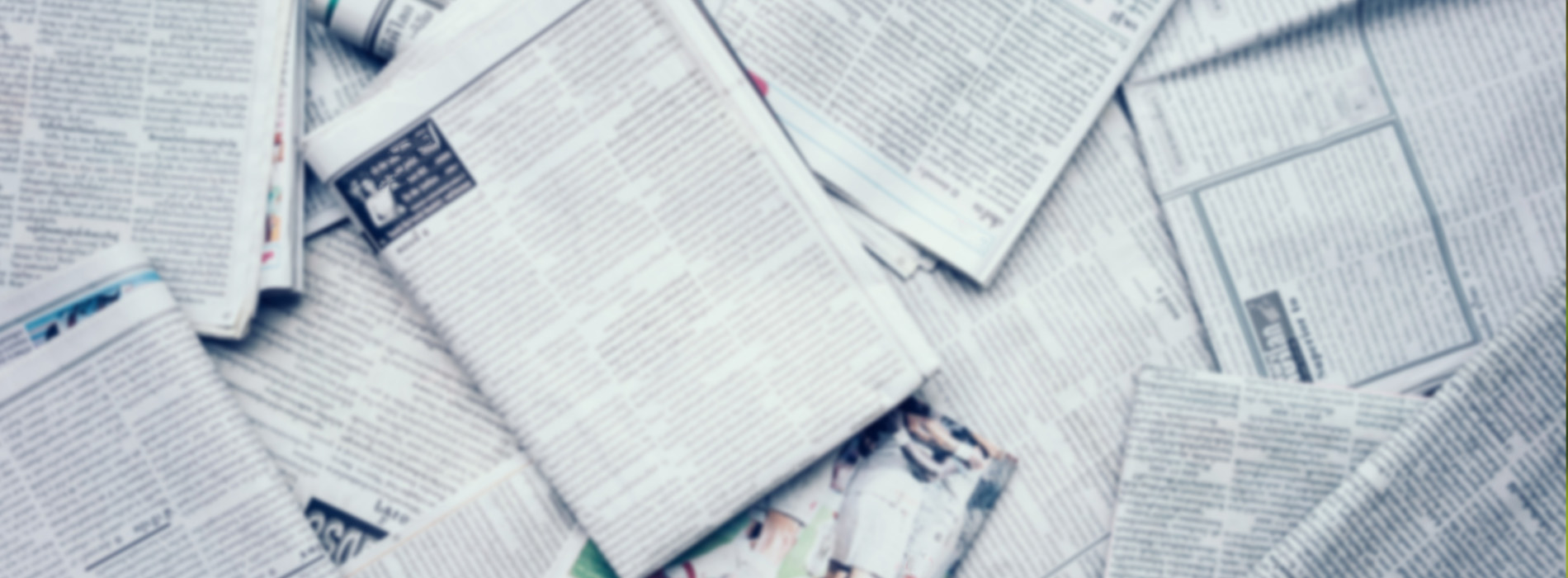 The image shows a collection of newspapers with visible text, piled together in a way that obscures individual articles, against a neutral background.
