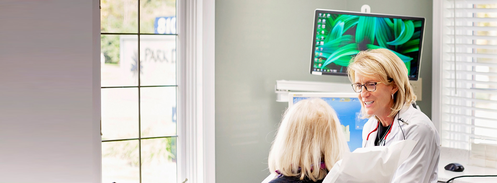 The image shows a person sitting at a desk with a computer monitor and keyboard, wearing glasses, and smiling towards the camera.