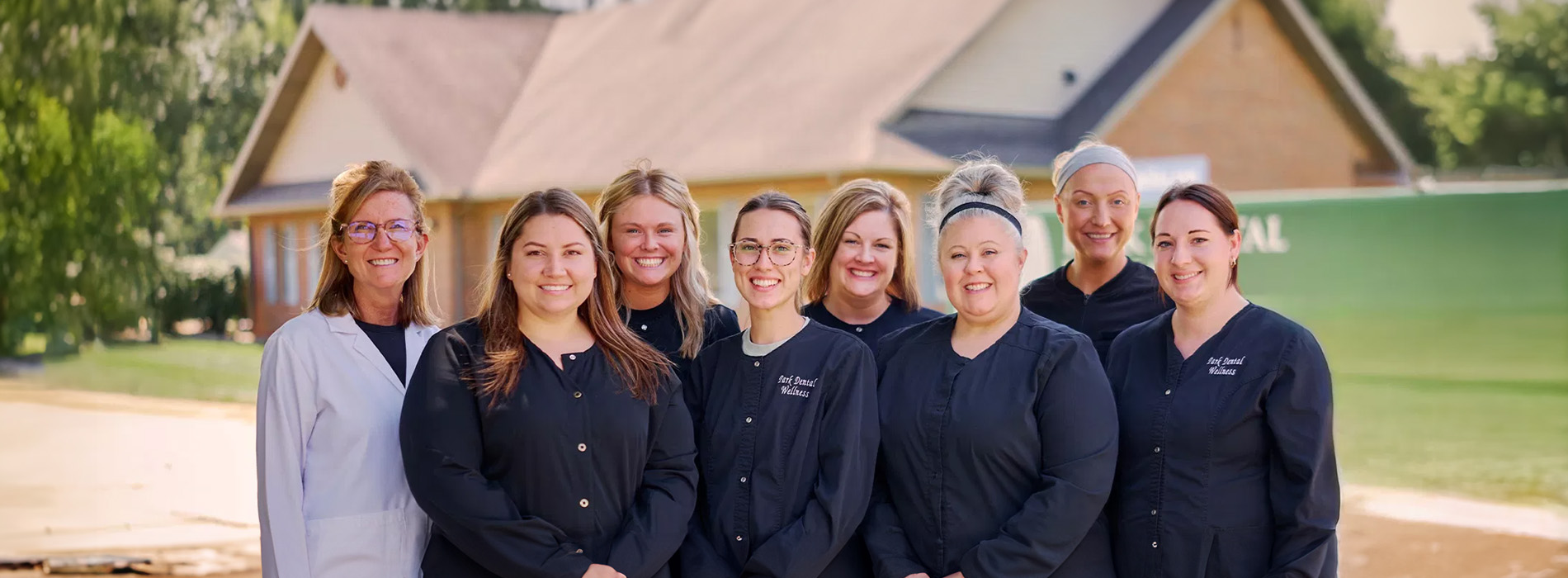 The image shows a group of seven individuals posing together outdoors, with a building in the background. They appear to be professionals, possibly healthcare workers, wearing name tags and standing behind each other.