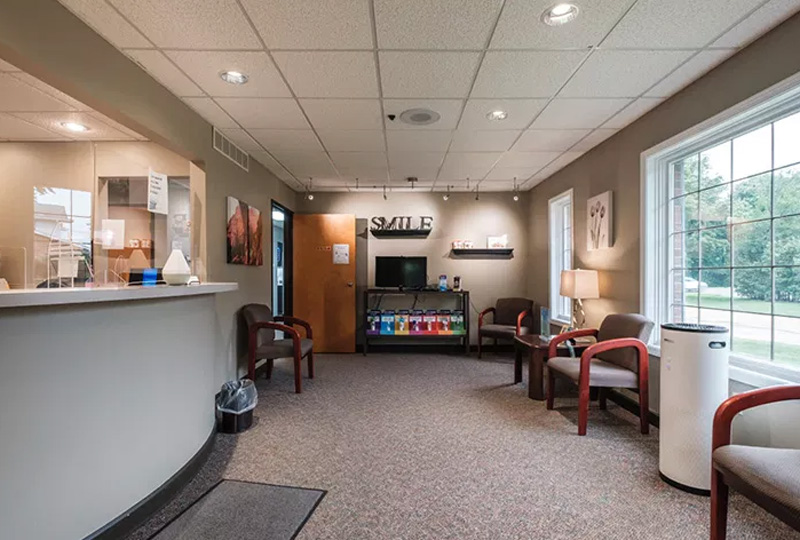 The image shows an interior view of a well-lit waiting room with a reception desk, chairs, and a television screen mounted on the wall.