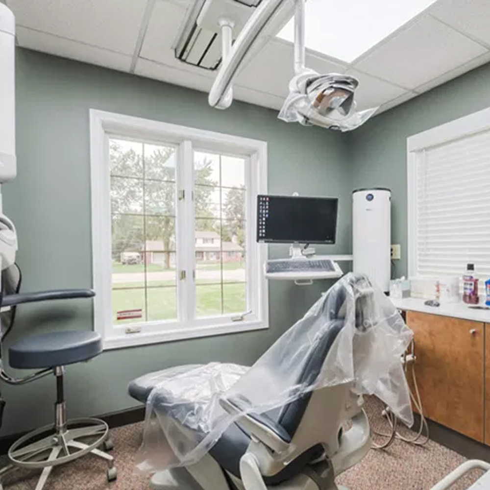 The image shows an interior view of a dental office with equipment such as a chair, monitor, and desk visible, along with dental instruments on a countertop and a window allowing natural light into the room.