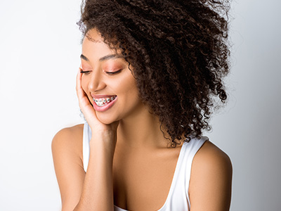 The image shows a woman with curly hair, wearing a white tank top, smiling at the camera with her hand on her face.