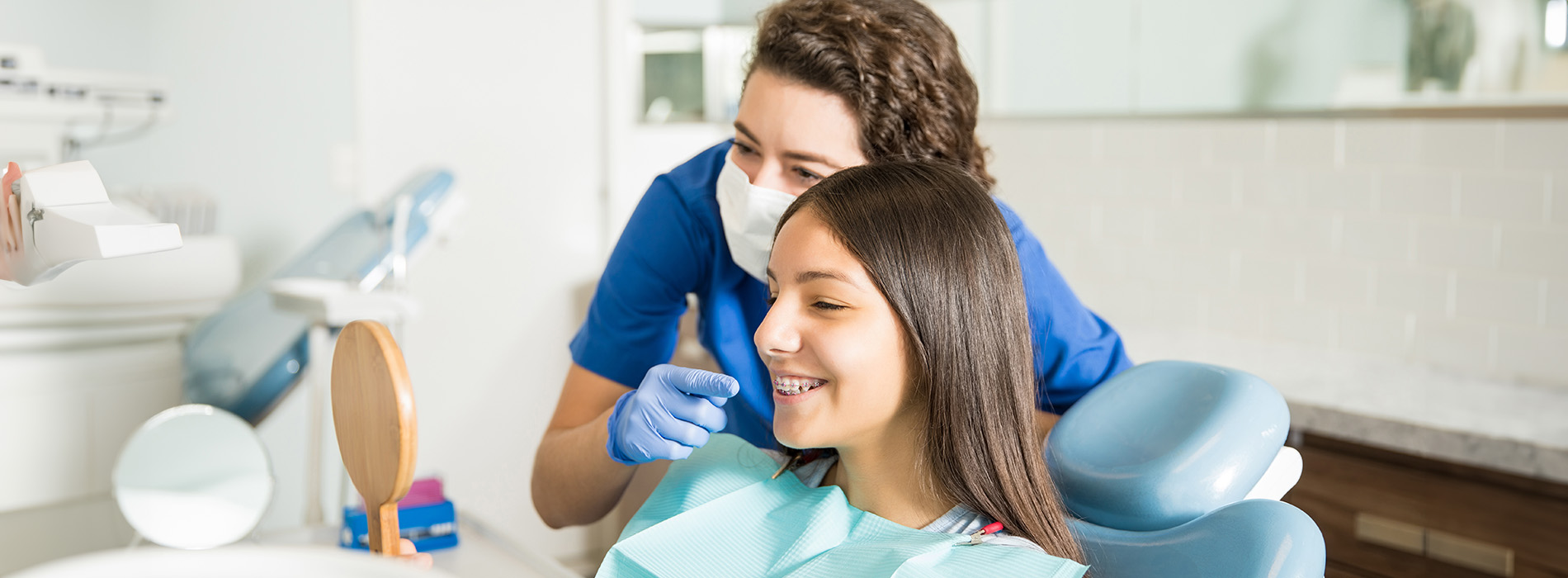 In the image, there are two individuals in a dental office setting  a woman is seated in a dental chair with her head resting on a dental device, while another person, presumably a dentist or dental hygienist, stands nearby holding a mirror and smiling at the camera.