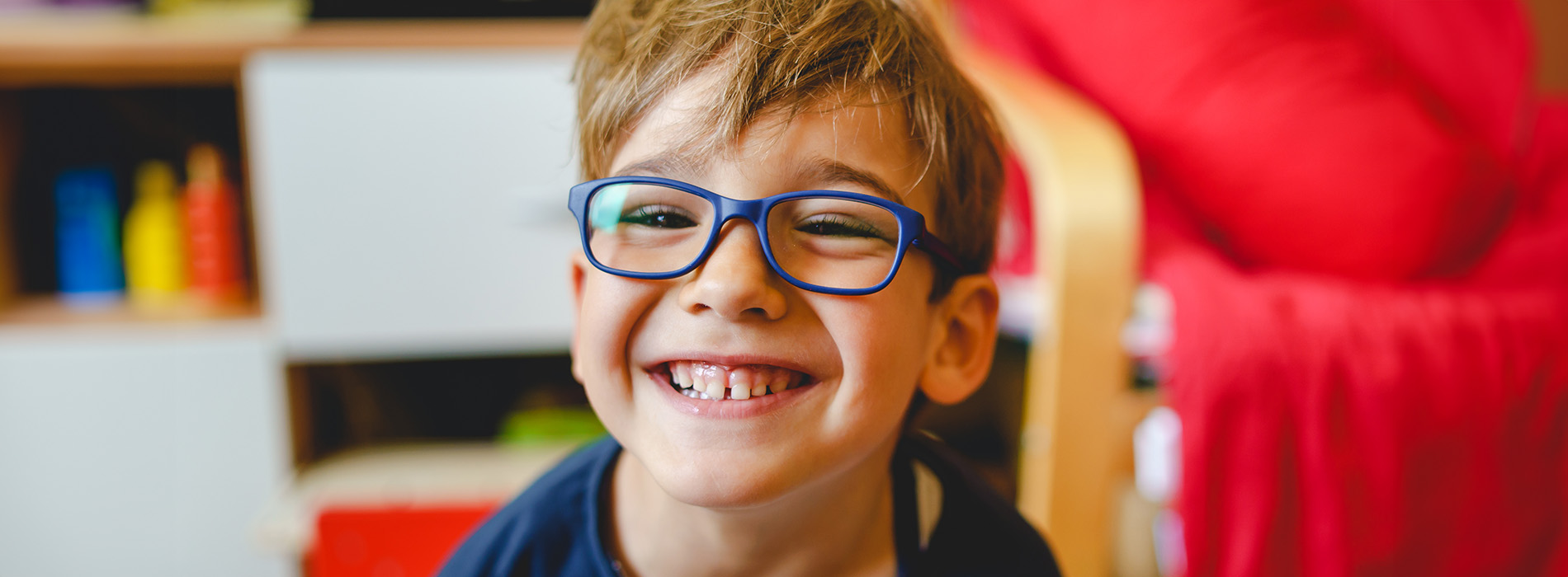 A young boy with glasses and blue eyes, smiling at the camera, wearing a dark shirt, sitting on a red chair with books behind him, in a room with white walls and a window.