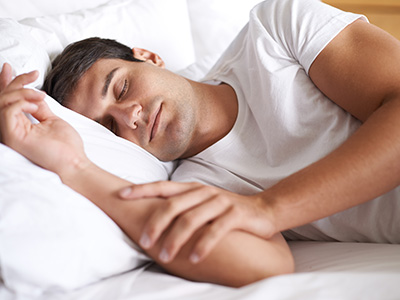 The image shows a man lying on his side with his head resting on one hand, appearing to be asleep. He is wearing a white t-shirt and dark pants, and there are no visible texts or distinguishing marks in the image.
