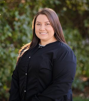 The image shows a woman standing outdoors with a smile, wearing a black jacket over a white shirt, posing against a background of greenery.