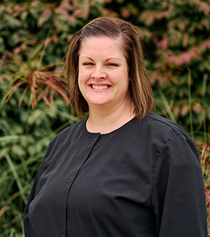 A smiling woman wearing a black shirt poses outdoors in front of a building with greenery.